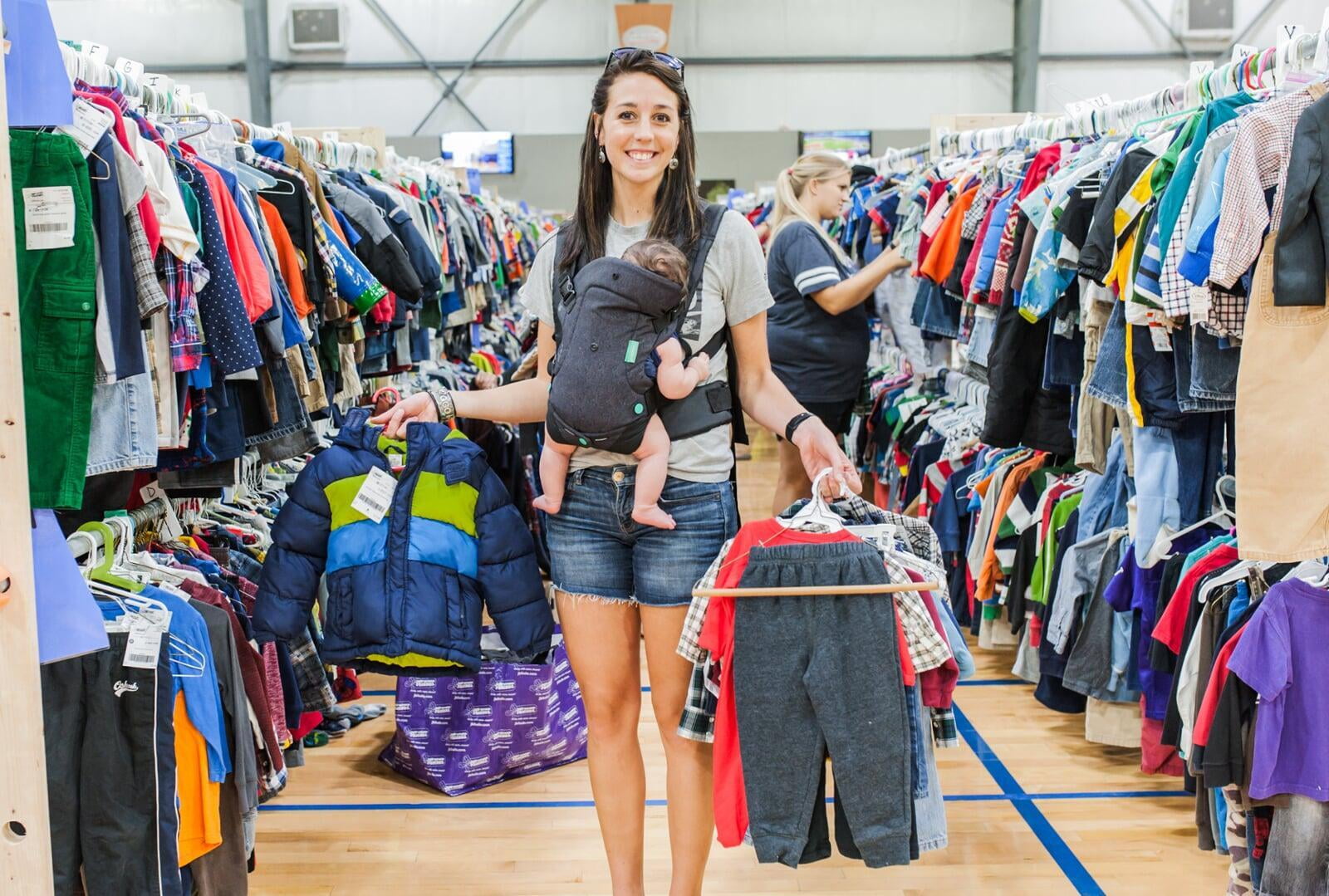 Mom wears baby and holds toddler boy clothes in each hand. Second mom shops behind her from the racks of boys clothes.