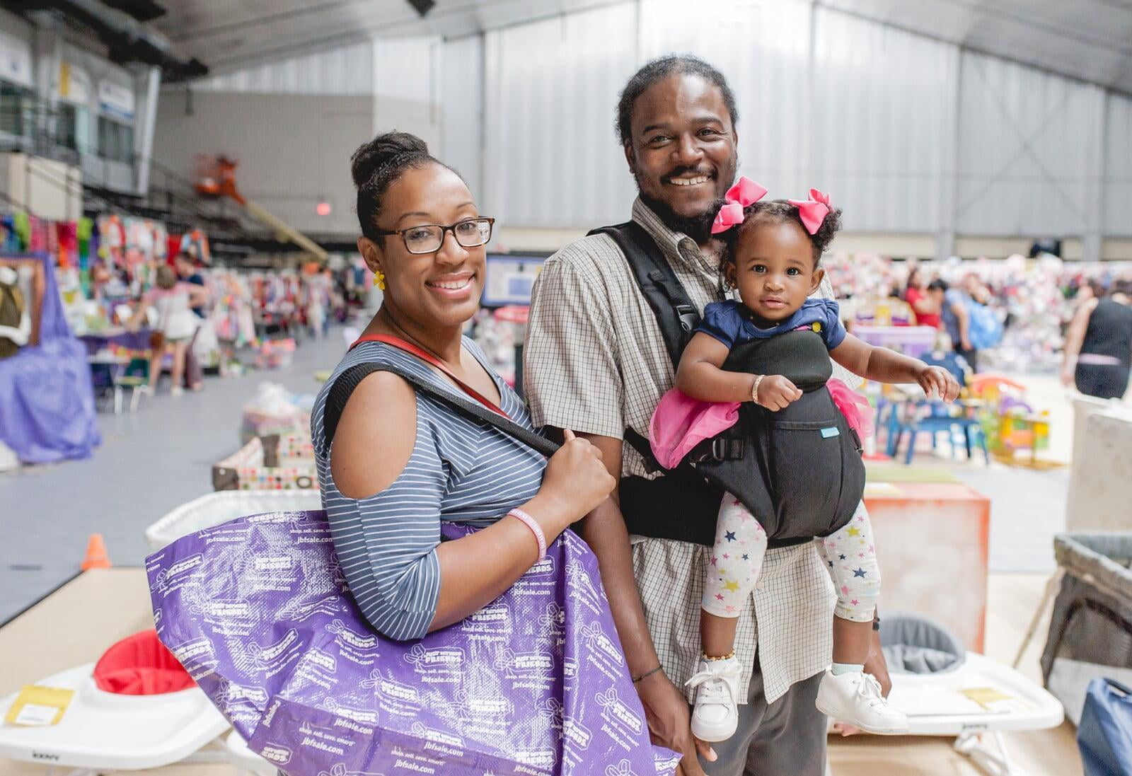 A mom and dad shopping at the JBF sale. Mom is holding a purple JBF shopping bag and dad carries his daughter in a front-pack carrier.