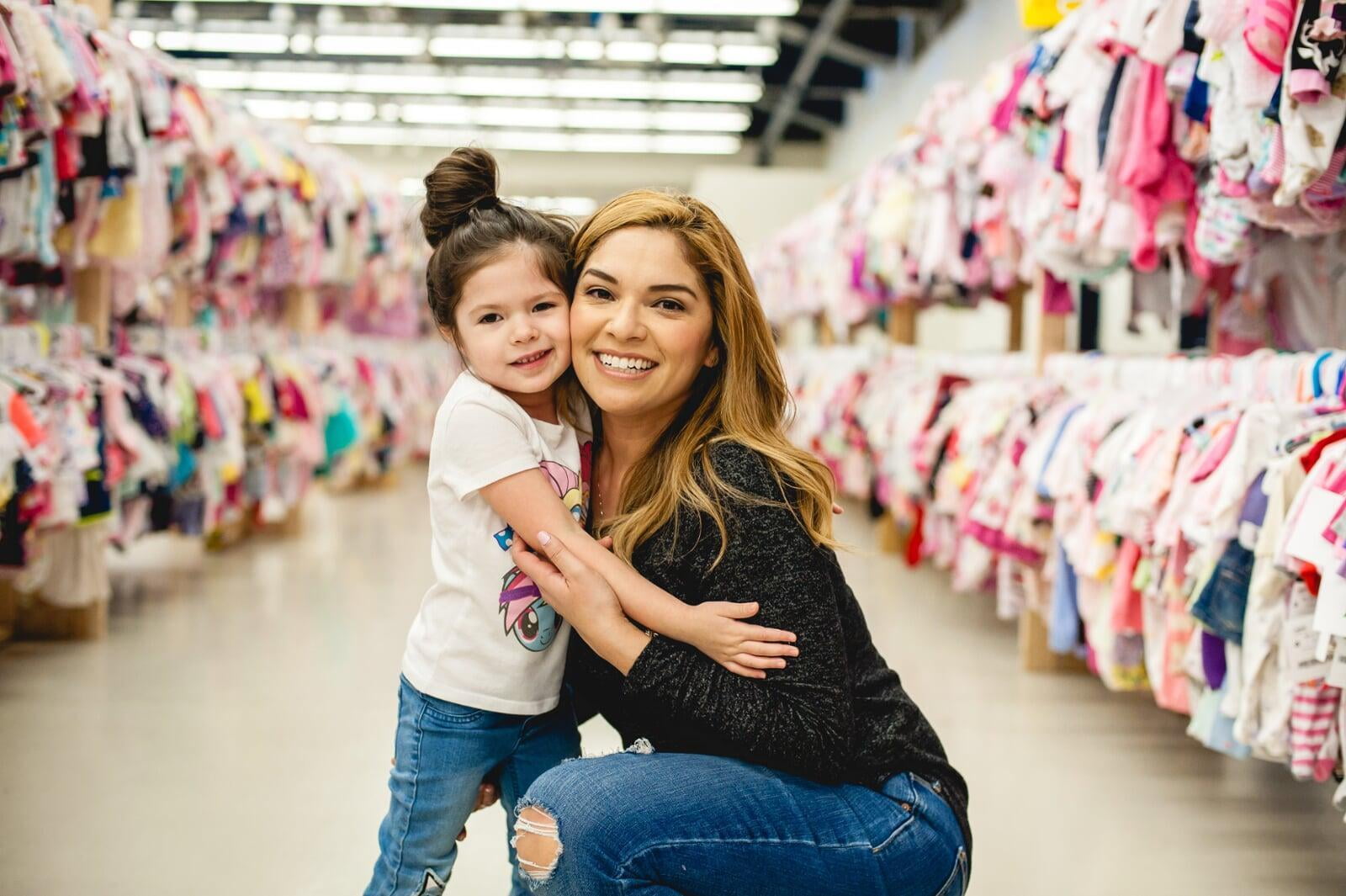 Mom smiles as she carries her baby in a front-pack carrier. She is walking past tables covered with items for sale at Just Between Friends.